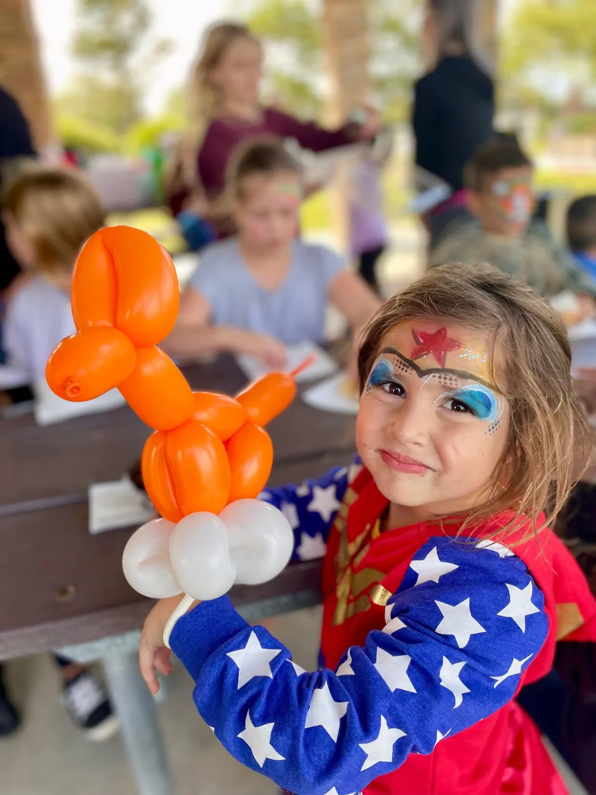 A girl in a superhero costume holding an orange balloon dog at an outdoor party table.