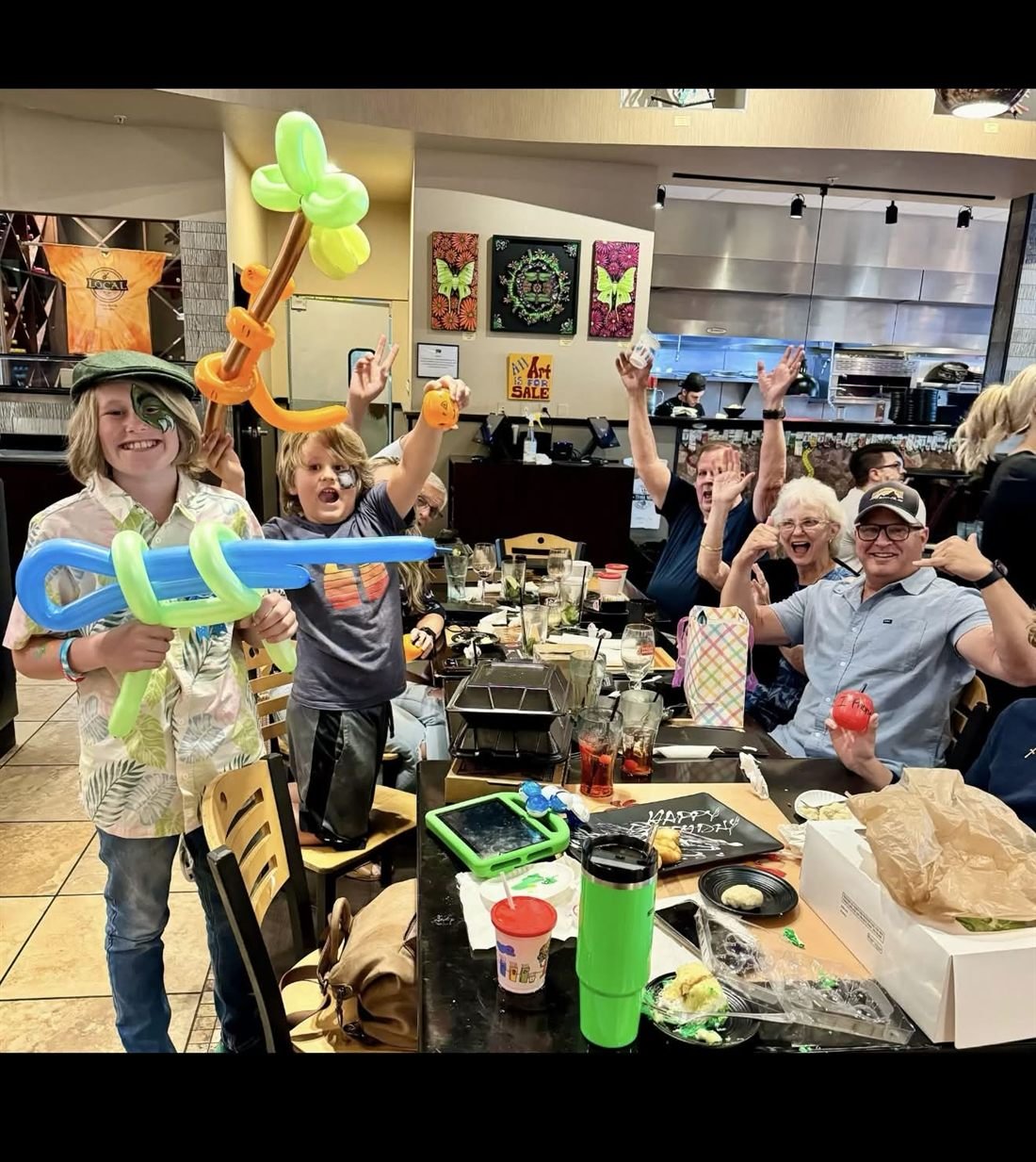 A restaurant birthday party. Two boys hold twisted balloon swords and a palm tree while the whole table cheers behind them.