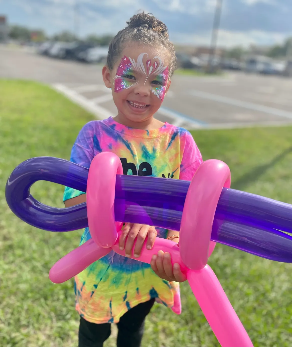A child in a tie-dye shirt holding a large pink and purple twisted balloon creation.