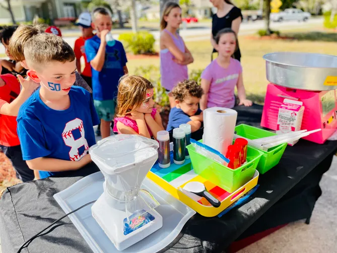 An outdoor concessions table with a cotton candy machine and Pucker Powder supplies while children wait in line.
