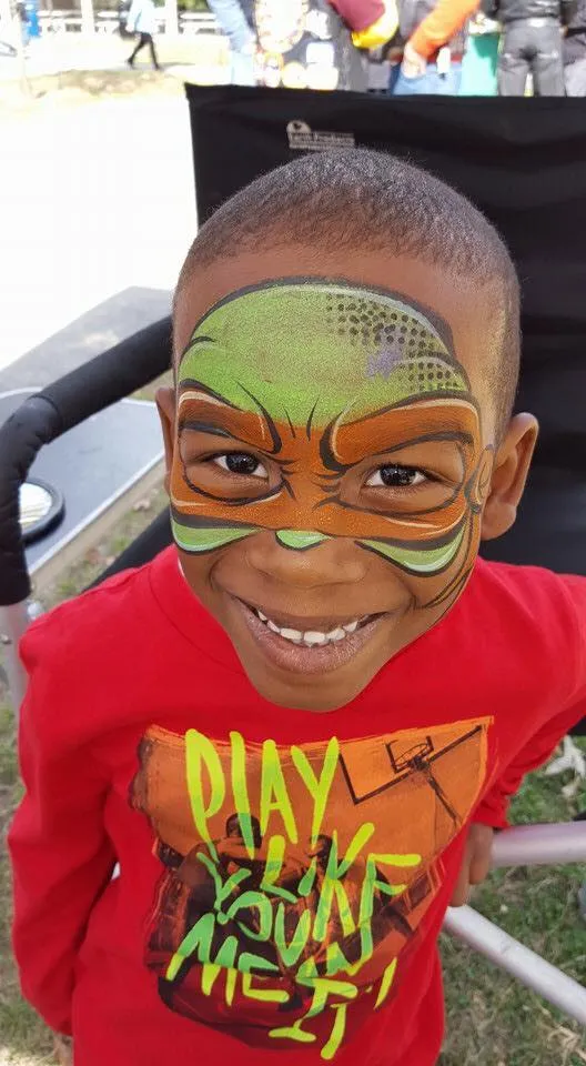 A grinning boy with a green and orange superhero-style mask painted across his eyes.