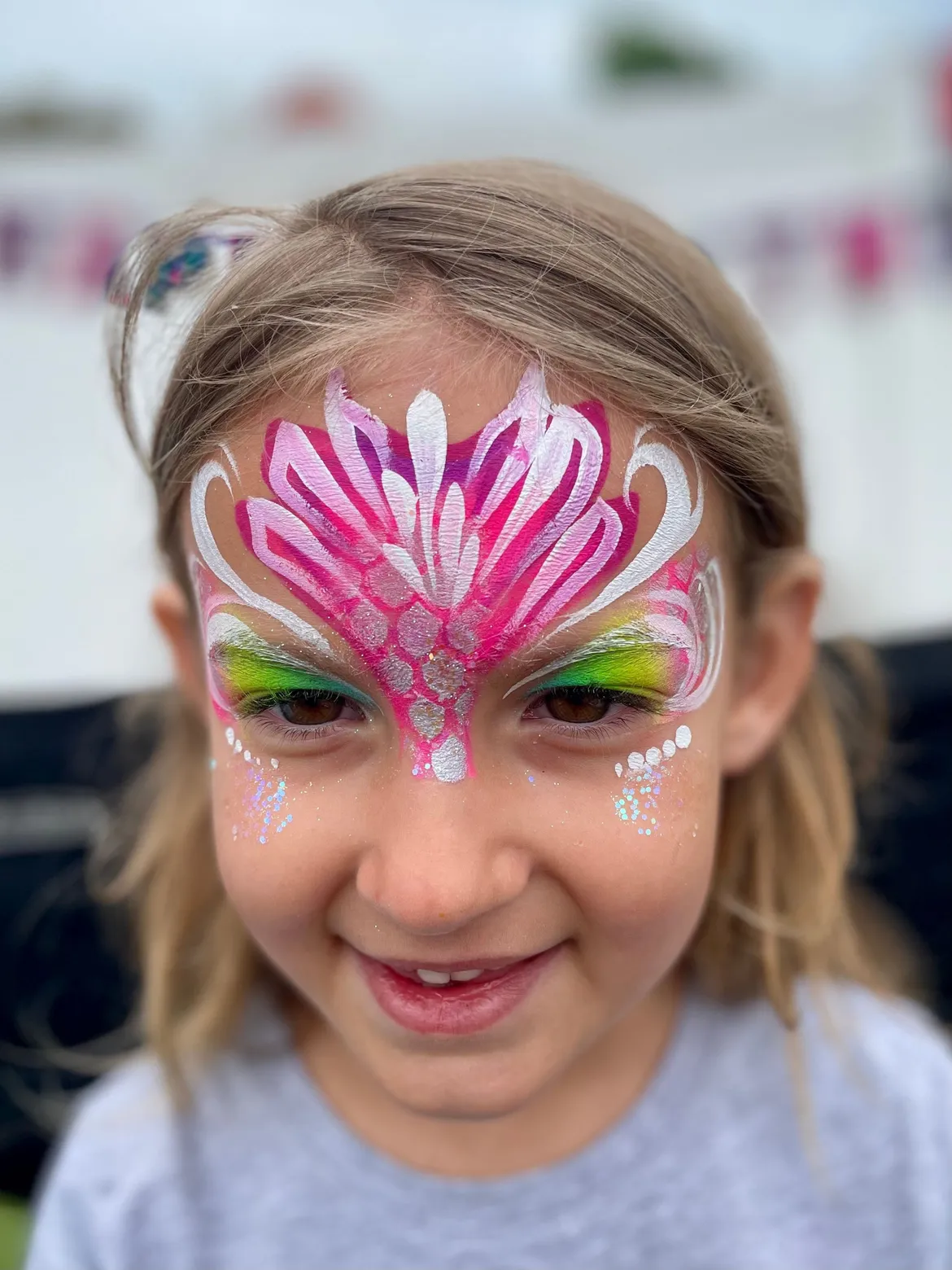 A smiling child wearing a pink and white crown design painted across their forehead and eyes.