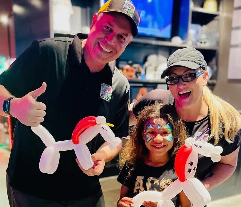 Two Cheerioograms performers in branded shirts holding balloon dogs beside a smiling girl with butterfly face paint.