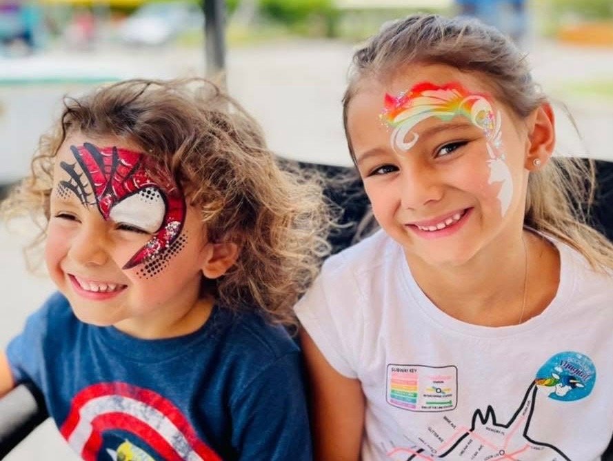 Two children smiling side by side — one with a red spider-hero mask painted across his eyes, the other with a rainbow and white swirl design across her forehead.