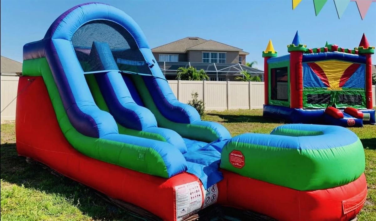 A tall blue and green inflatable water slide with a splash pool, beside a castle bounce house on a sunny backyard lawn.